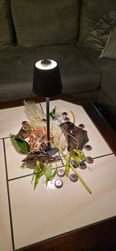 Auf einem Couchtisch steht eine Lampe mit Kronkorken und Pflanzendekoration um sie herum verstreut.