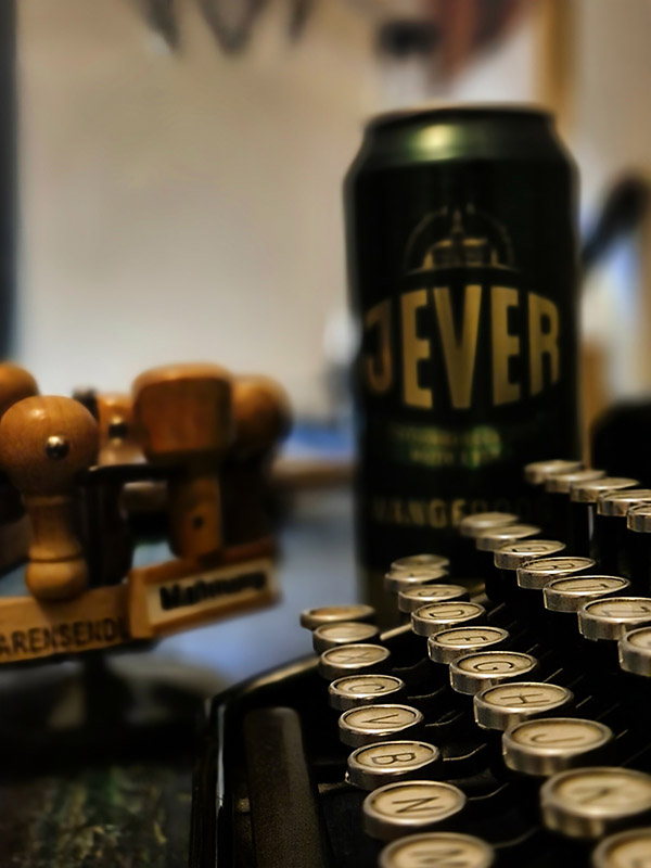 Hinter einer Schreibmaschine steht eine Jever-Bierdose. Links neben der Dose ist ein Stempelträger mit Stempeln.