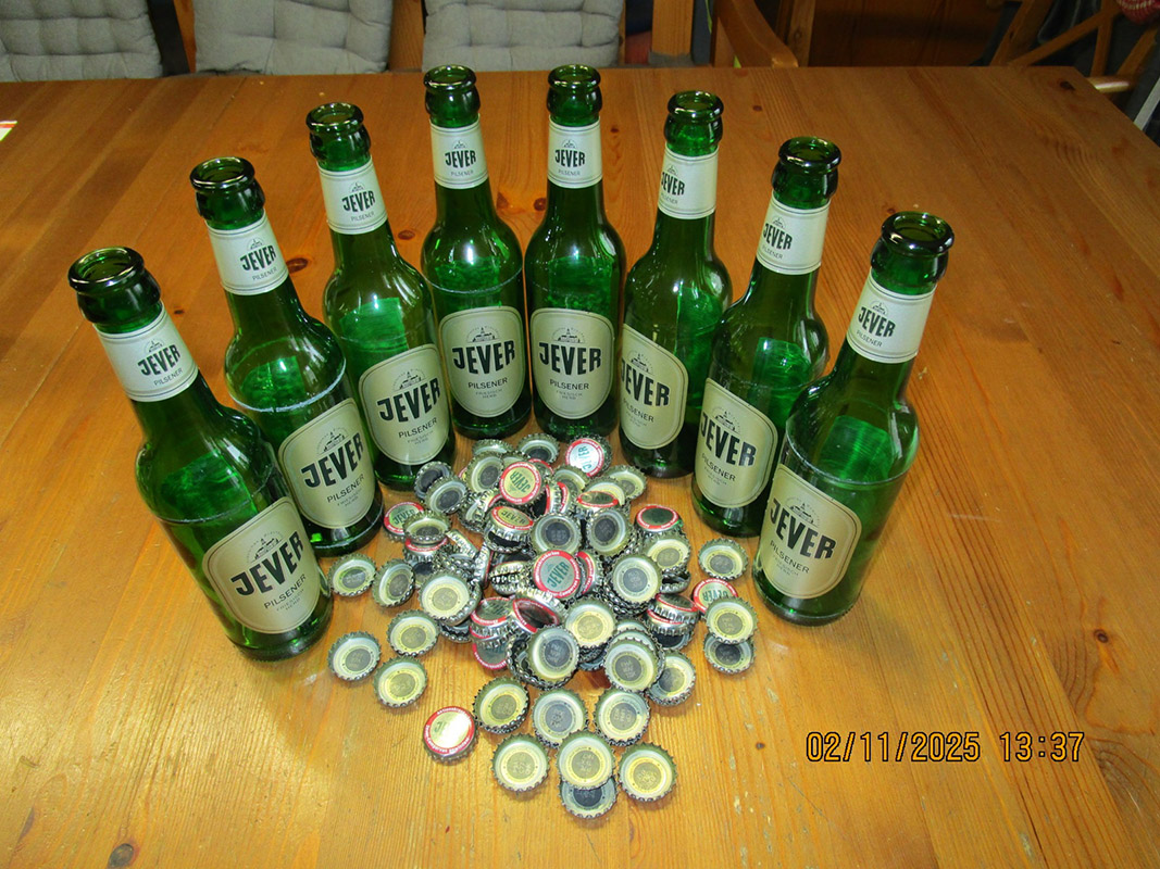 Mehrere Jever-Bierflaschen stehen im Halbkreis nebeneinander. Vor ihnen liegt ein Haufen Kronkorken.