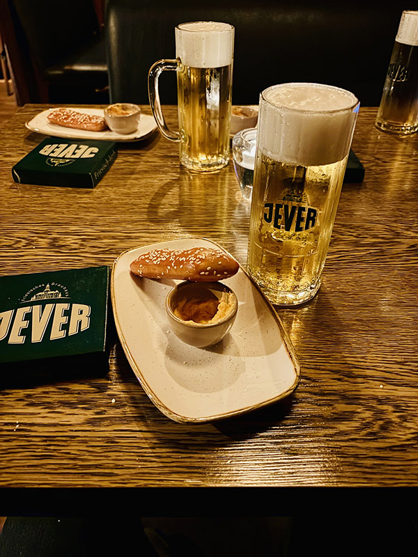 Auf einem Esstisch stehen gegenüber voneinander jeweils ein Glas Bier mit Jever-Aufschrift, eine Laugenstange mit Dip und ein Jever-Deckel.