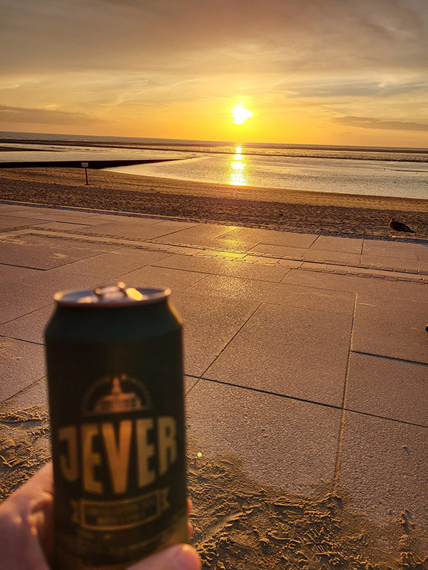 Jemand hält eine Jever-Bierdose in der Hand. Im Hintergrund ist ein Strand. Die Sonne färbt den Himmbel gelb und orange.