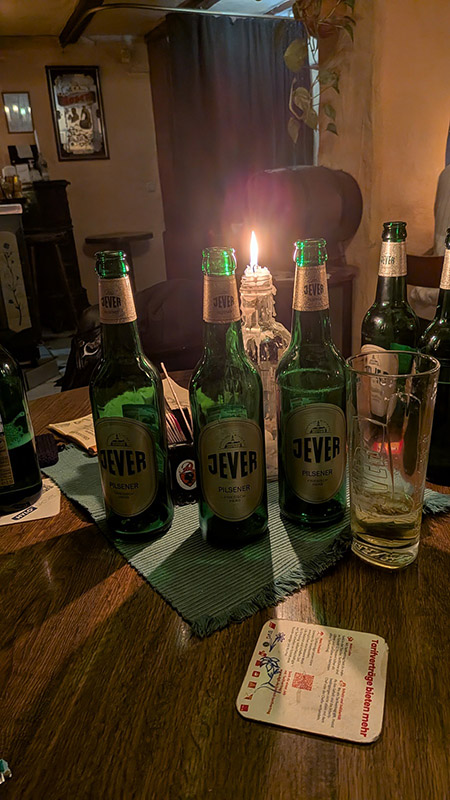 Mehrere Jever-Bierflaschen stehen nebeneinander auf einem Tisch. Hinter ihnen beleuchtet eine Glasflasche mit einer brennnden Kerze darin den dunklen Raum.