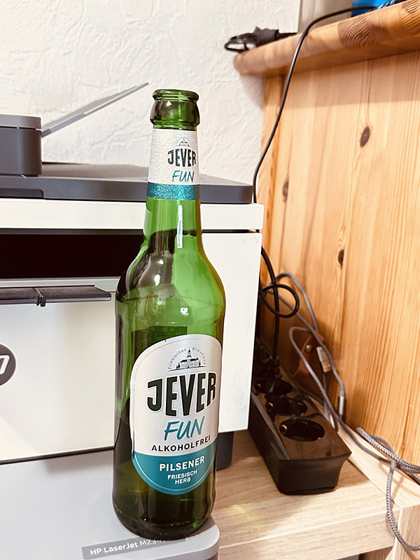 Eine Jever-Bierflasche steht auf einem Drucker. Rechts daneben ist eine Multisteckdose mit Kabeln.