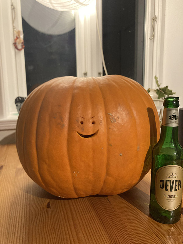 Ein Kürbis mit einem kleinen, geschnitzten Gesicht liegt links neben einer Jever-Bierflasche.
