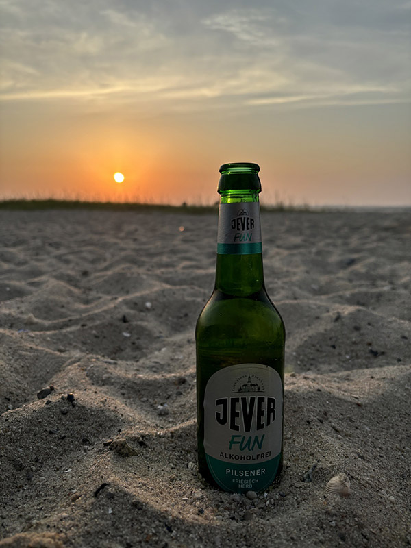 Eine Jever-Bierflasche steht im Sand. Im Hintergrund färbt die Sonne den Himmel orange.