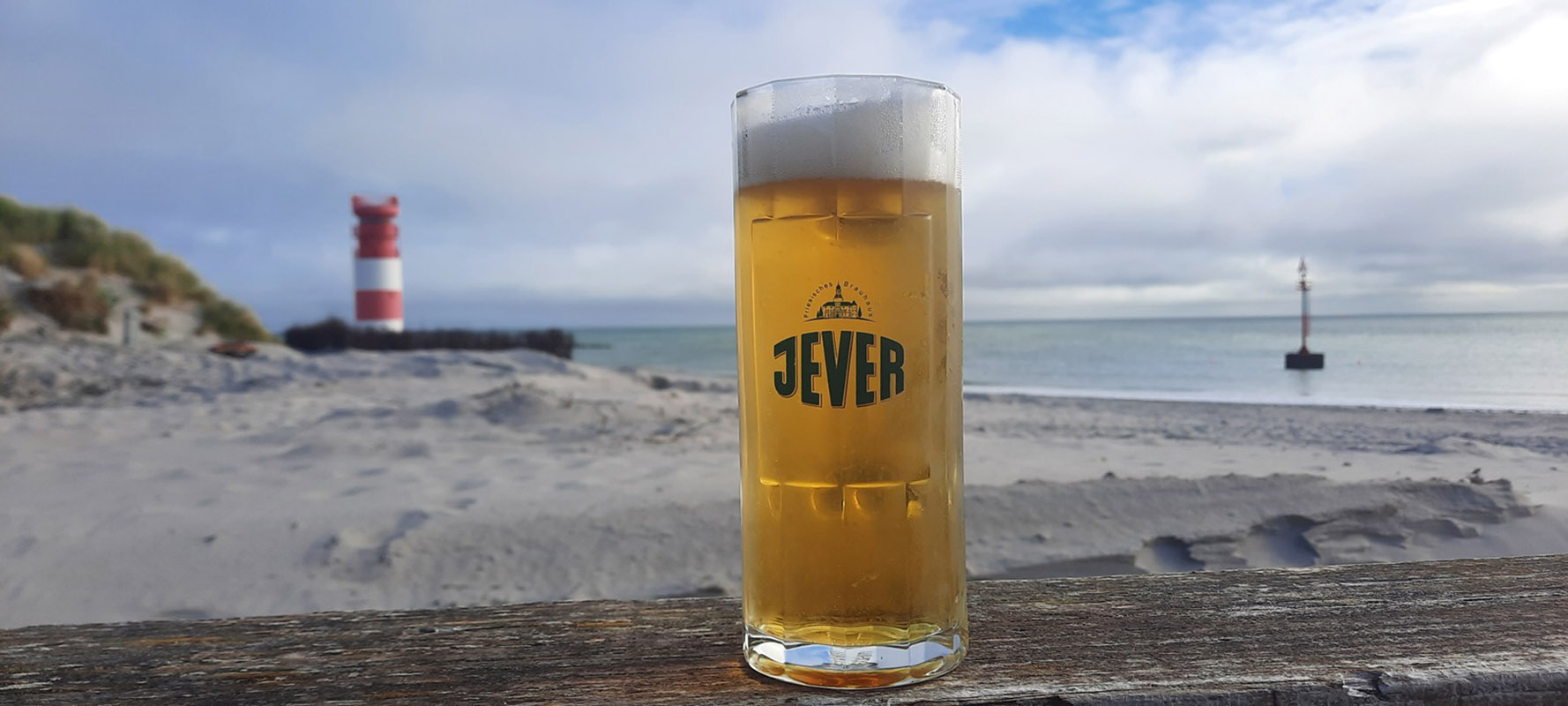 Ein Jever-Glas steht auf einem Geländer. Im Hintergrund ist ein Strand mit einer Düne und einem rot-weiß gestreiften Leuchtturm.