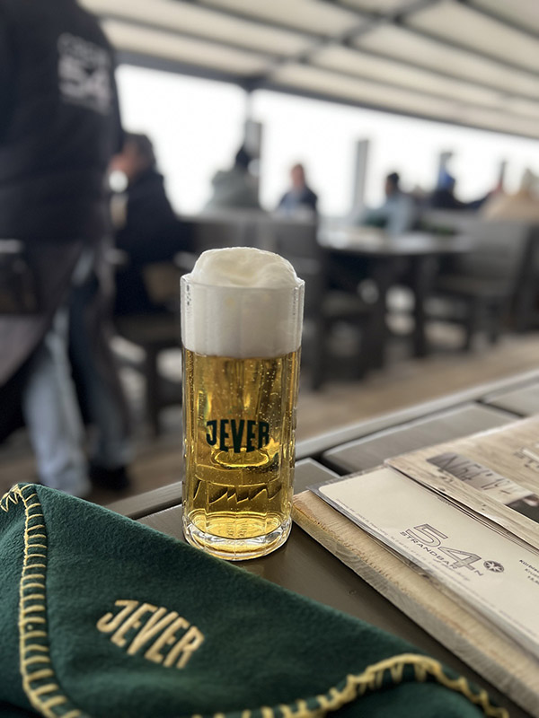 Ein Jever-Glas mit Bier darin steht auf einem Tisch. Daneben liegt eine Jever-Decke. Im Hintergrund sitzen mehrere Personen an Tischen.