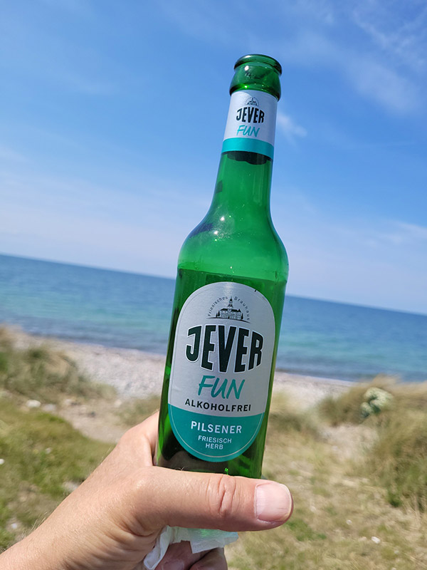 Jemand hält eine Jever-Bierflasche hoch. Im Hintergrund ist ein Strand mit einer Düne.