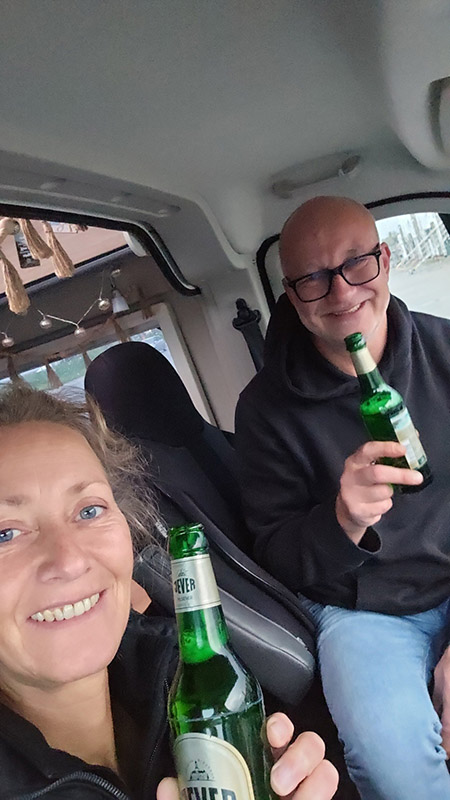 Eine Frau und ein Mann sitzen nebeneinander in einem Auto und halten jeweils eine Jever-Bierflasche hoch.