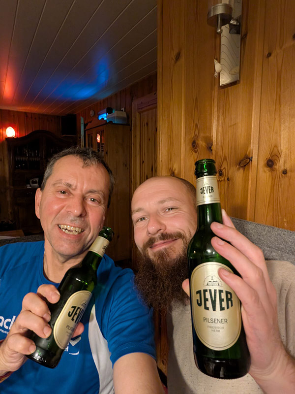 Zwei Männer sitzen nebeneinander und halten lächelnd jeweils eine Jever-Bierflasche hoch.