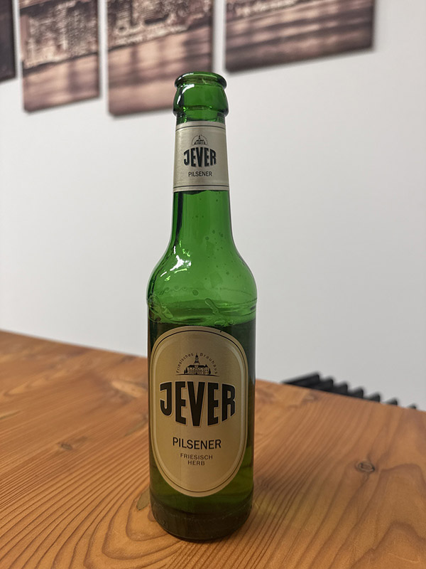 Eine Jever-Bierflasche steht auf einem Tisch. Im Hintergrund hängen Bilder an der Wand.