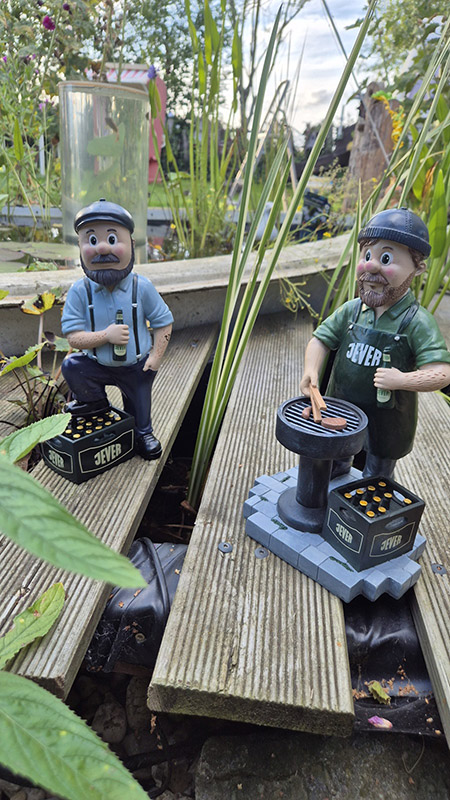 Auf dem Holzbrettboden in einem Garten stehen zwei Figuren von jeweils einem Mann mit einem Jever-Bierkasten.