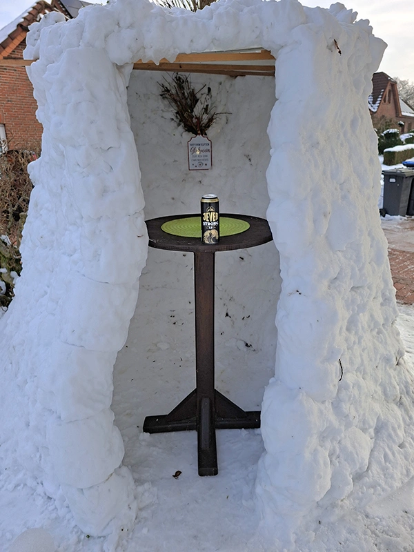 Kleines Iglu aus Schnee mit Tisch im Inneren, darauf eine Getränkedose Jever. Im Hintergrund Häuser in einem Wohngebiet.