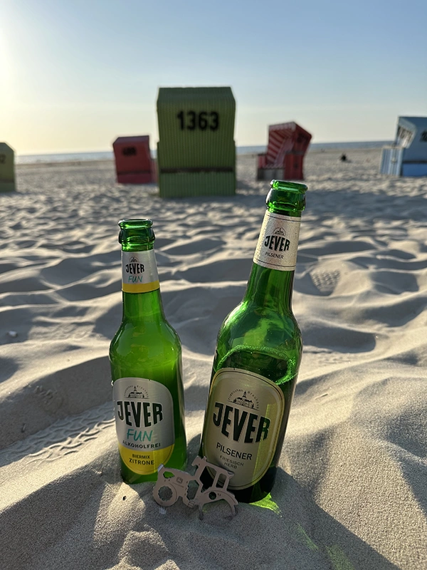Zwei Jever-Flaschen stehen im Sand am Strand, mit bunten Strandkörben im Hintergrund bei Sonnenuntergang.