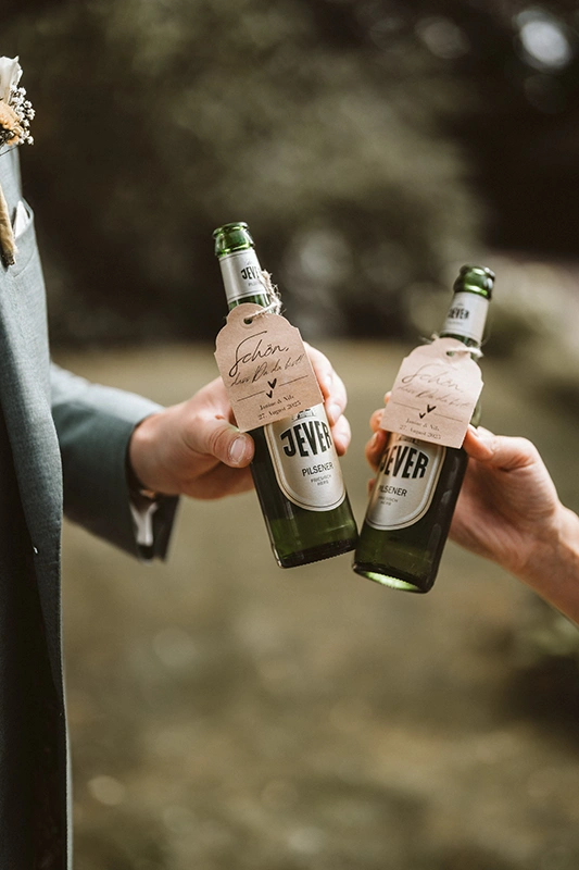 Zwei Personen stoßen mit Flaschen Jever Pilsener an. Im Hintergrund unscharfe Umgebung, an den Flaschen hängen Etiketten.