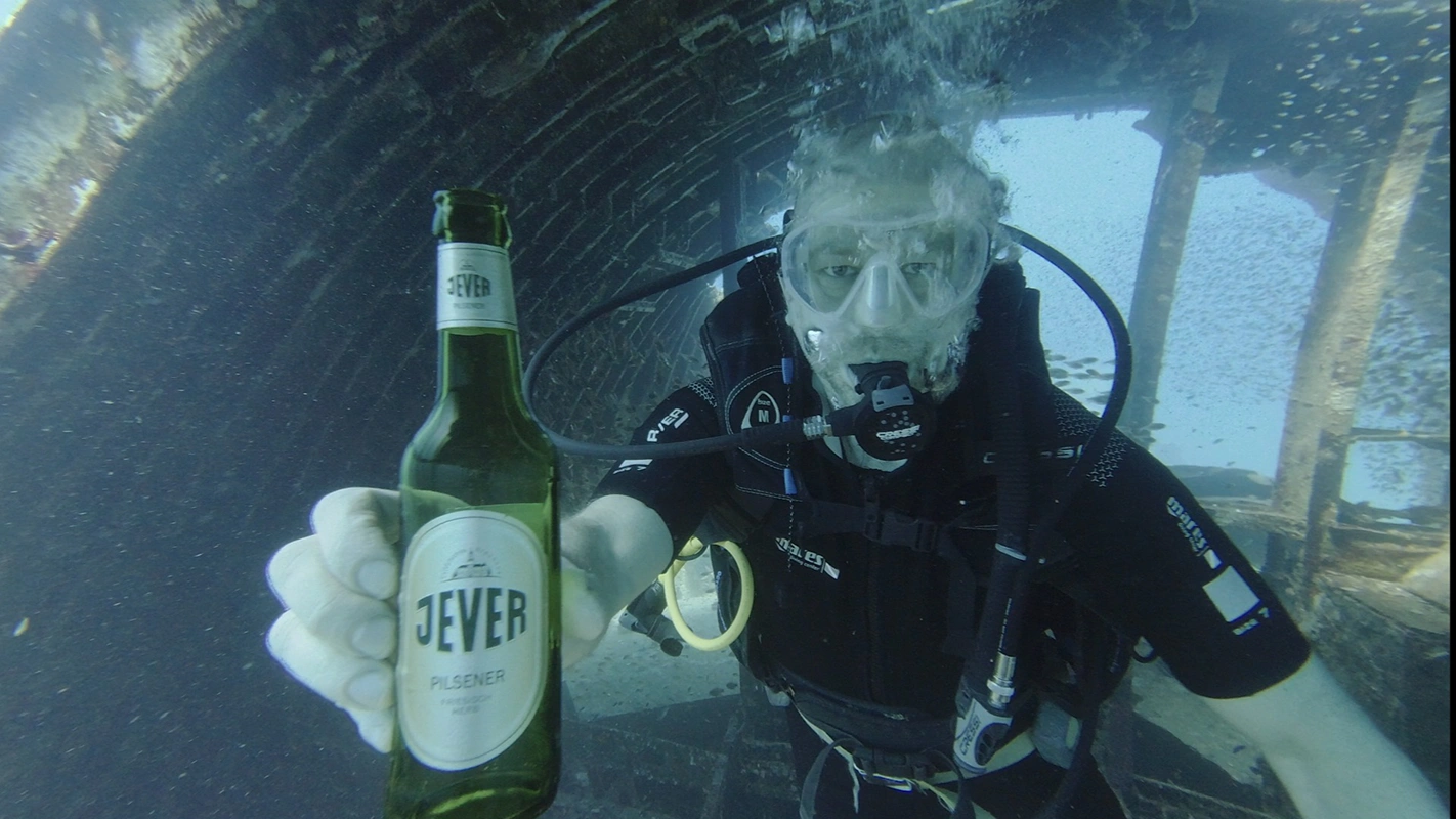 Taucher unter Wasser hält eine Flasche Jever Pilsener in der Hand. Im Hintergrund Wrack, Luftblasen und Fische.