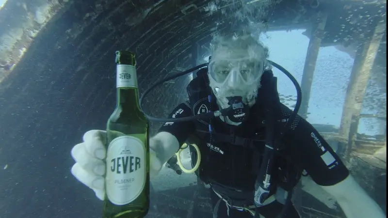 Taucher unter Wasser hält eine Flasche Jever Pilsener in der Hand. Im Hintergrund Wrack, Luftblasen und Fische.