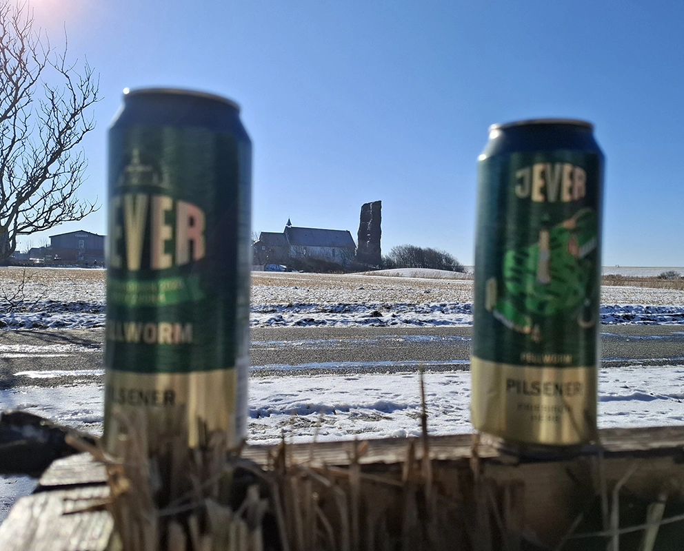Zwei Jever Bierdosen auf einem Holzzaun. Im Hintergrund verschneite Landschaft mit Gebäude.