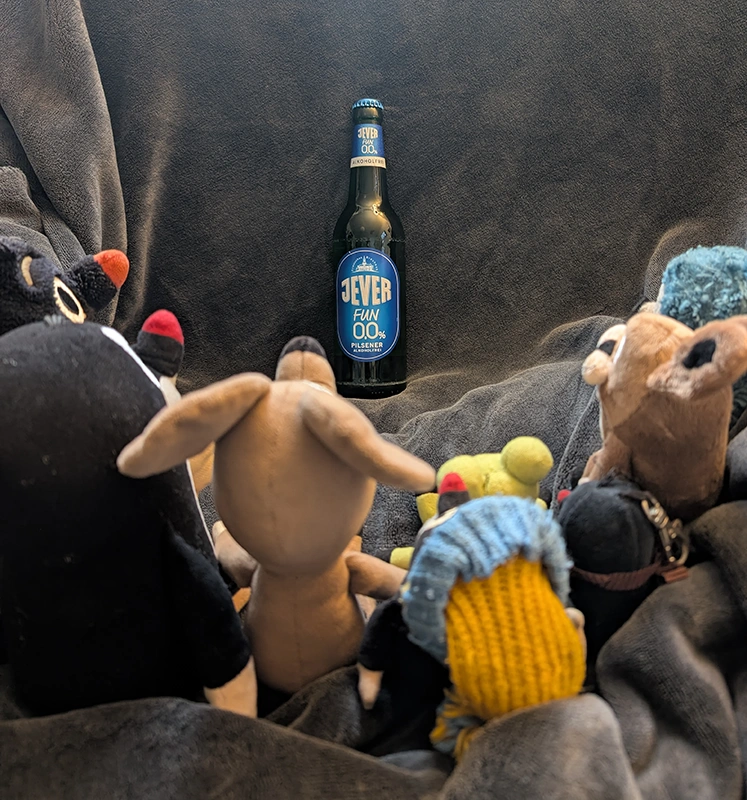 Eine Gruppe Plüschtiere sitzen um eine Flasche Jever Fun 0,0 % Alkoholfrei auf einer Decke.