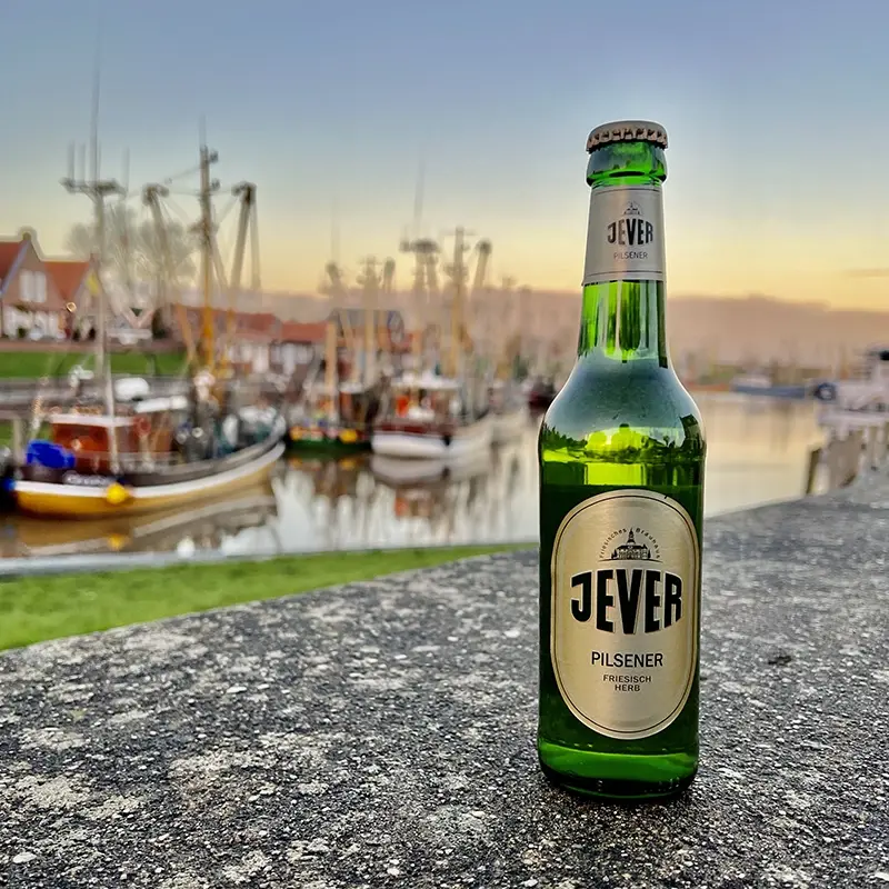 Jever Bierflasche auf einer Mauer. Im Hintergrund Hafen mit Fischerbooten bei Sonnenuntergang.
