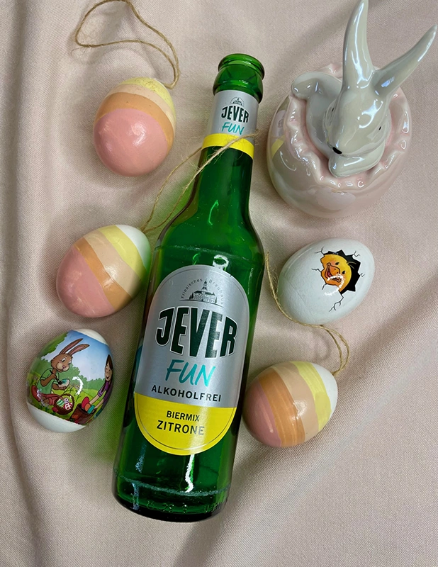 Jever Fun Zitrone Bierflasche umgeben von bunten Ostereiern und einer weißen Hasenfigur auf rosa Stoff.