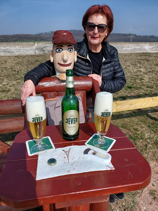Eine Frau legt ihre Arme um eine Holzfigur. Vor ihnen ein Tisch mit Jever Bierflasche, zwei Jever Gläsern und Bierdeckel.