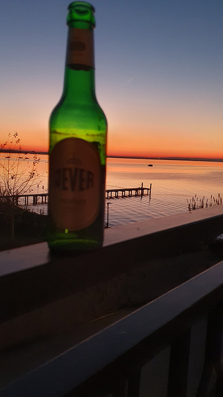Auf einem Geländer steht eine Jever-Bierflasche. Im Hintergrund ist ein Gewässer mit einem Steg. Der Himmel ist zum Teil orange und rot gefärbt.