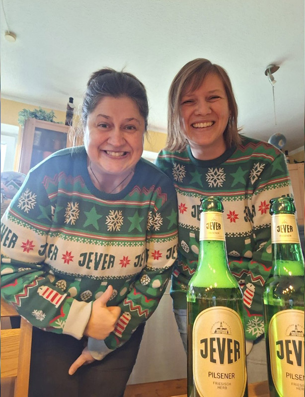 Zwei Frauen in Jever-Weihnachtspullovern stehen lächelnd hinter zwei Jever-Bierflaschen.