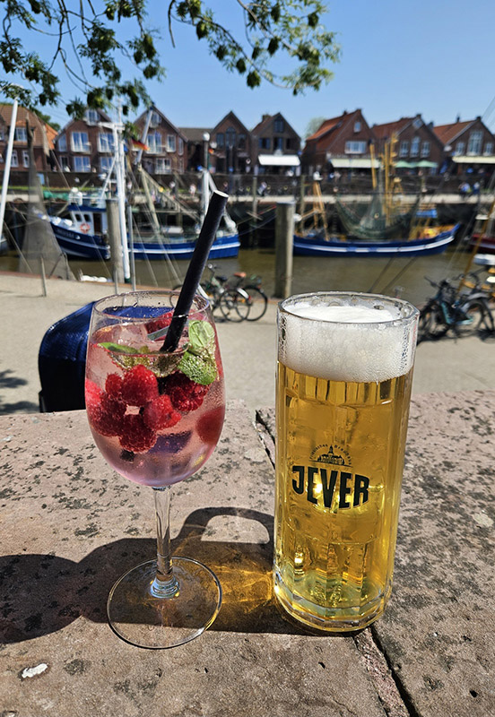 Ein Glas mit Bier und Jever-Aufschrift steht rechts neben einem Cocktail. Im Hintergrund ist ein Hafen mit Booten.