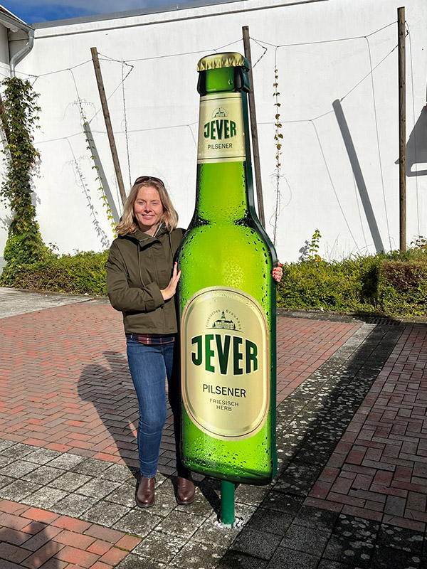 Eine Frau steht neben einem großen Aufsteller einer Jever-Bierflasche.