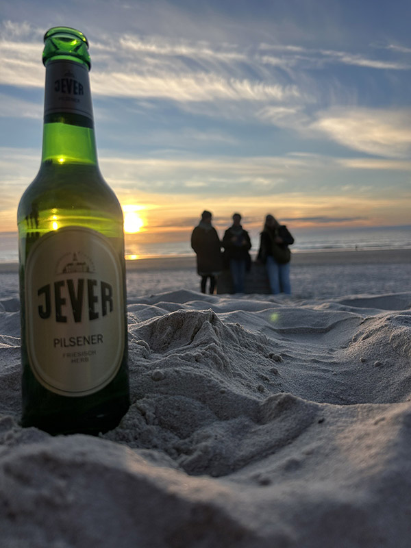 Eine Jever-Bierflasche steht im Sand eines Strandes. Im Hintergrund steht eine Gruppe Personen. Die Sonne färbt den Himmel orange.