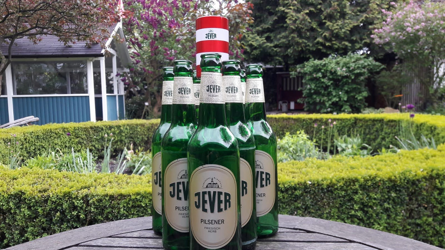Mehrere Jever-Bierflaschen sind wie Bowlingkegel auf einem Tisch angeordnet. Dahinter steht ein kleiner Jever-Leuchtturm. Im Hintergrund ist ein Garten.