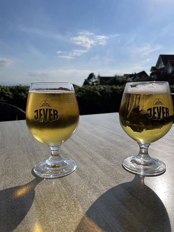 Zwi Gläser mit Jever-Aufschrift und Bier darin stehen nebeneinander auf einem Tisch. Im Hintergrund sind Häuser und eine Hecke.