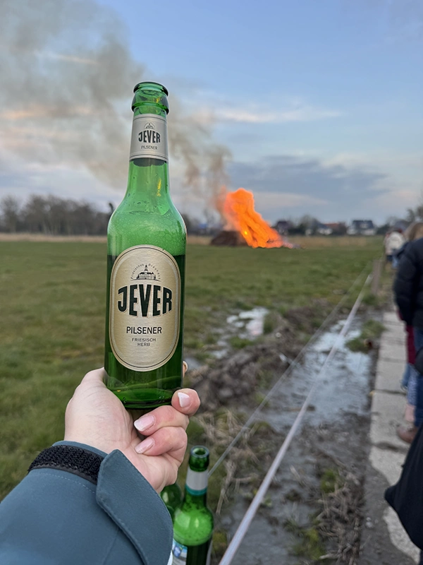 Eine Hand hält eine Jever-Bierflasche im Vordergrund, während im Hintergrund ein großes Feuer brennt.