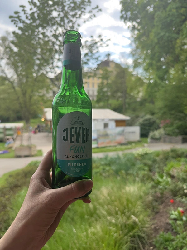 Eine Hand hält eine grüne Flasche Jever Fun alkoholfrei im Freien, mit Bäumen und einem Gebäude im Hintergrund.