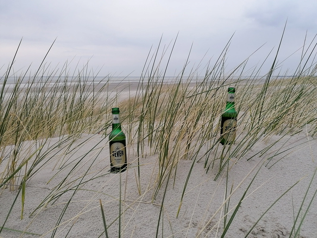Jever Bierflaschen im Sand, umgeben von Dünengras. Der Himmel ist bedeckt, und das Meer erstreckt sich im Hintergrund.