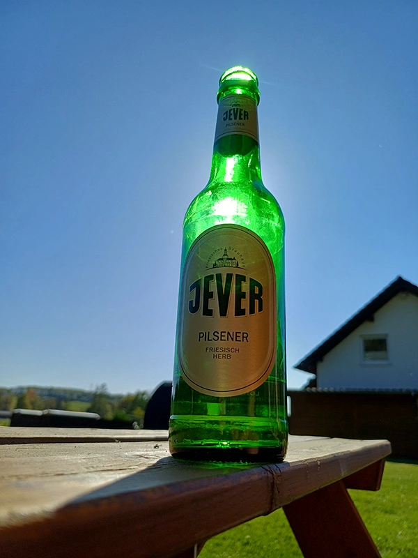 Eine Jever Pilsener Bierflasche steht auf einem Holztisch im Freien, beleuchtet von der Sonne. Ein Haus im Hintergrund.