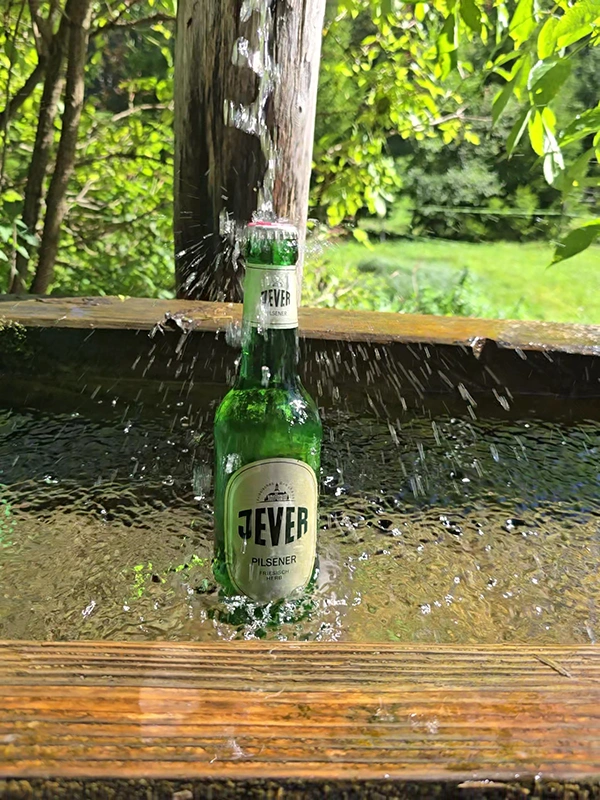 Eine Jever Bierflasche steht unter einem Wasserstrahl in einem hölzernen Brunnen, umgeben von grüner Natur im Hintergrund.