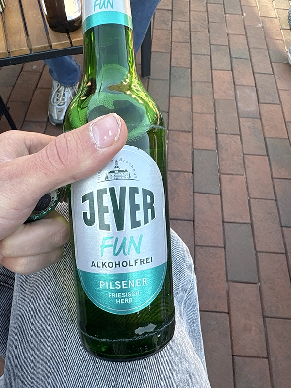 Person hält eine Flasche der Marke Jever Fun Alkoholfrei in der Hand. Der Hintergrund zeigt eine gepflasterte Fläche.
