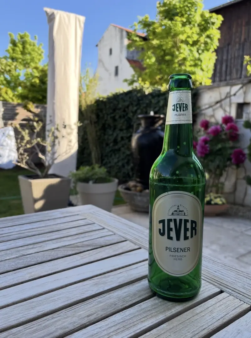 Jever Bierflasche auf einem Holztisch im Freien. Im Hintergrund blühende Pflanzen und ein altes Gebäude bei Sonnenschein.