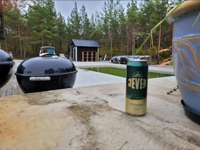 Bierdose von  Jever Pilsener steht auf einer Betonfläche neben Blumentopf und Grill. Im Hintergrund Bäume und ein Pool.