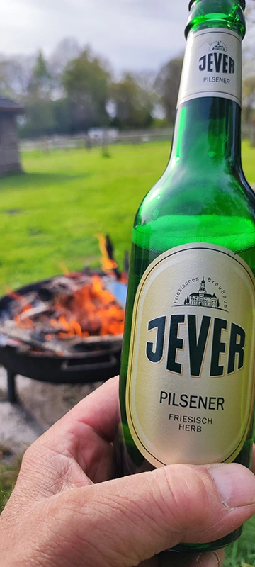 Person hält eine Jever Pilsener Flasche vor einem brennenden Lagerfeuer im Freien. Im Hintergrund eine grüne Wiese.