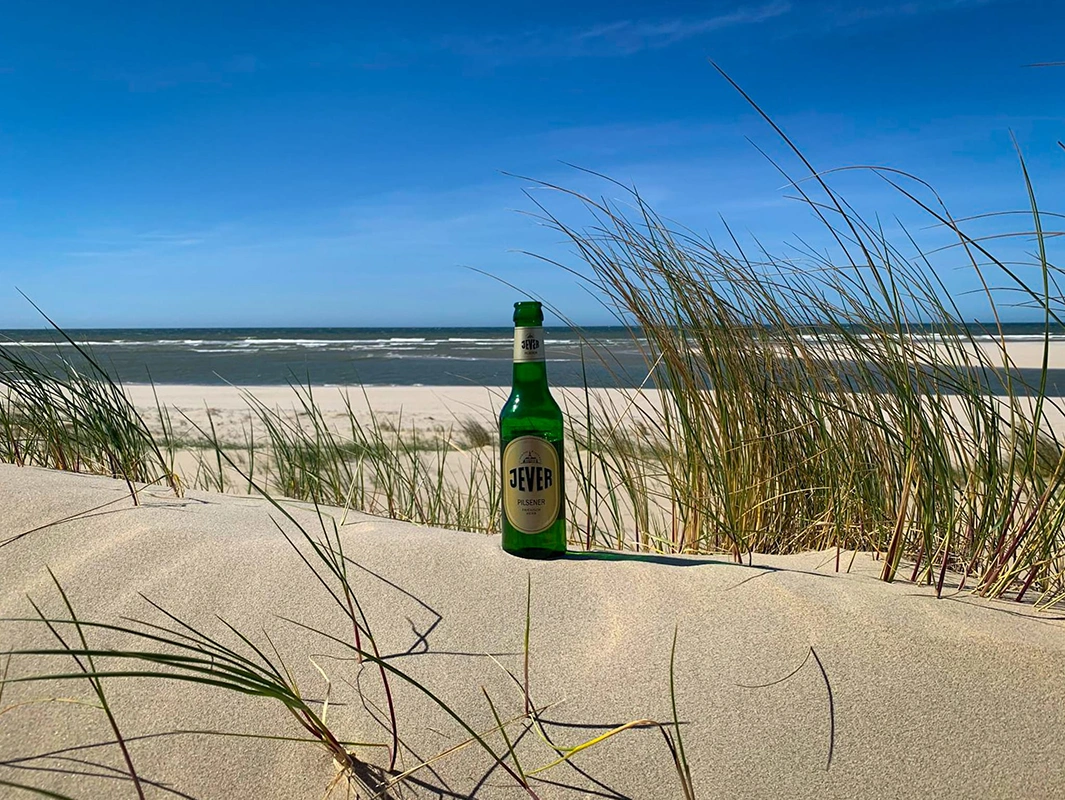 Eine Bierflasche von Jever steht im Sand einer Düne, umgeben von Gras. Im Hintergrund das Meer unter klarem, blauem Himmel.