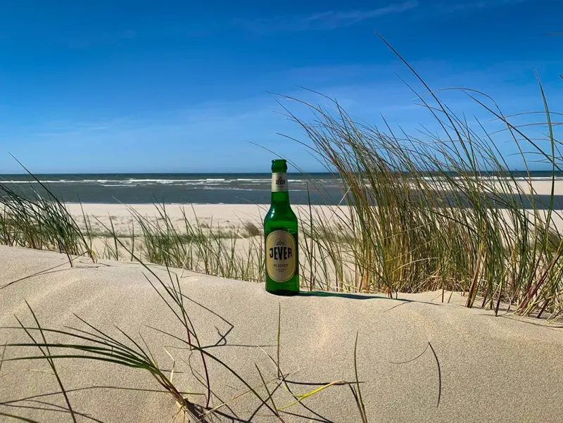 Eine Bierflasche von Jever steht im Sand einer Düne, umgeben von Gras. Im Hintergrund das Meer unter klarem, blauem Himmel.
