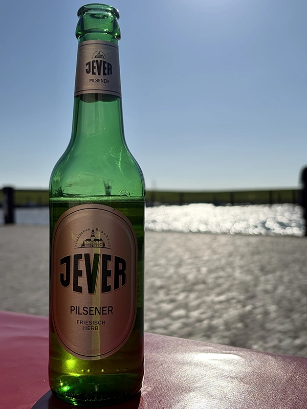 Jever Pilsener Flasche auf einem Tisch, mit einem sonnigen Himmel und Wasser im Hintergrund. 