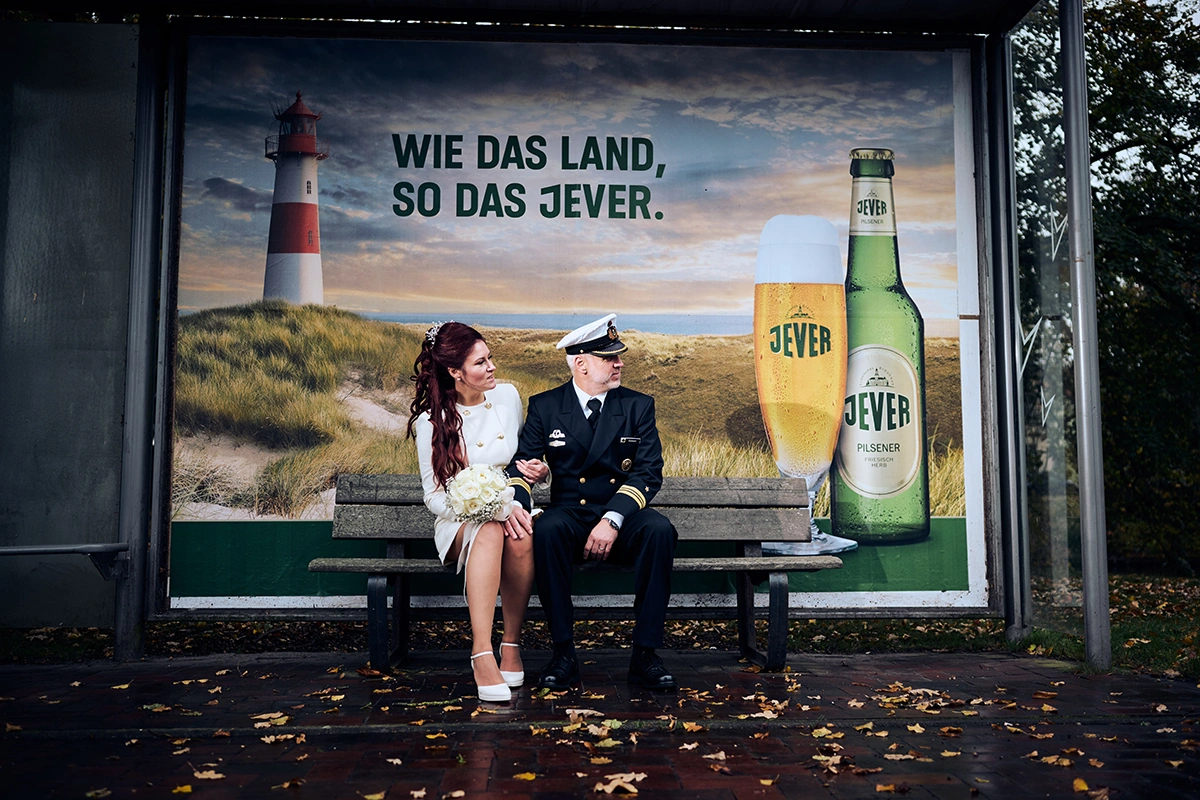 Ein Paar in Hochzeitskleidung sitzt auf einer Bank vor einem Jever-Werbeplakat mit Leuchtturm und Bierflasche im Hintergrund.
