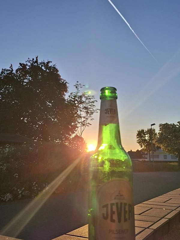 Jever Bierflasche im Vordergrund, während die Sonne im Hintergrund untergeht. Ein Kondensstreifen zieht sich über den Himmel.