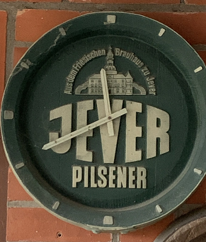 Runde Wanduhr mit Jever-Logo und Schriftzug Aus dem Friesischen Brauhaus zu Jever vor einer Backsteinwand.