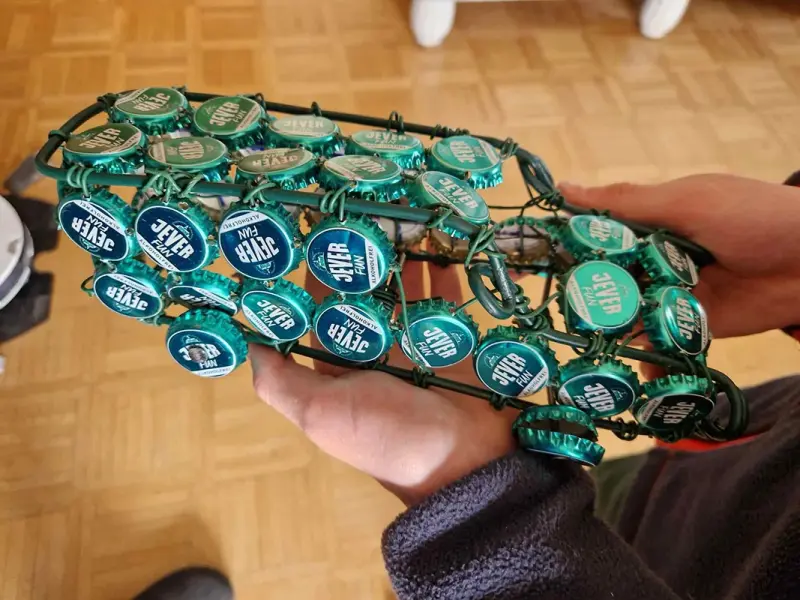 Ein handgefertigtes Auto aus Jever-Kronkorken wird von einer Person präsentiert. Der Boden ist aus Holzparkett.