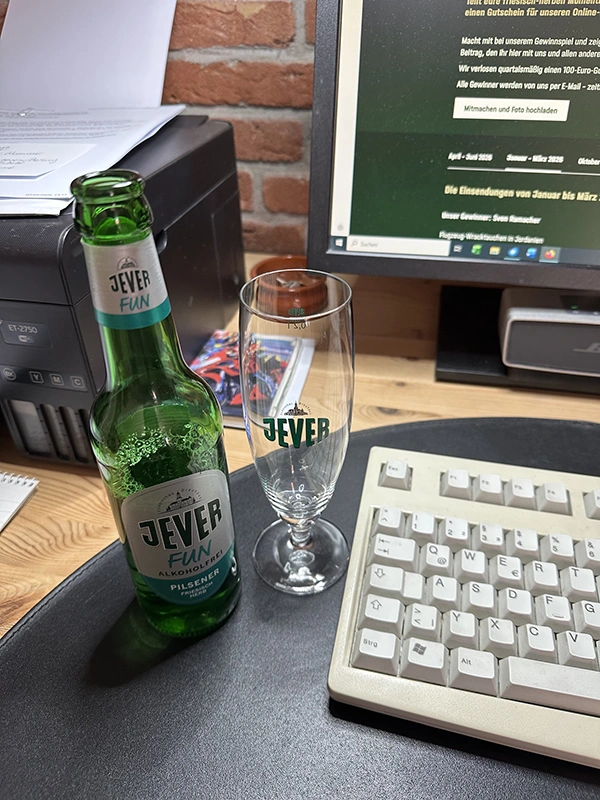 Eine geöffnete Flasche Jever Fun und ein leeres Jever-Glas stehen neben einer Tastatur auf einem Schreibtisch.